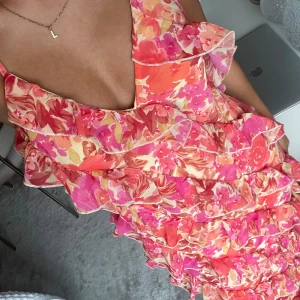 Blommig klänning med volanger - En färgglad långklänning med blommigt mönster i rosa, orange och vita nyanser. Klänningen har volanger längs hela längden och en fin v ringning