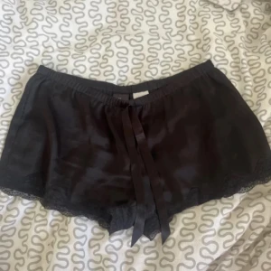 Svarta shorts med spetsdetaljer - Ett par svarta shorts med spetsdetaljer och en knytning framtill. Shortsen har en lös passform och är tillverkade i ett glansigt material.