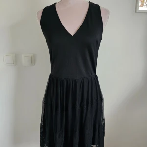 Svart ärmlös klänning - En elegant svart ärmlös klänning med v-ringning. Klänningen har en detaljerad spetskjol med broderade mönster och en lätt genomskinlig nederdel.