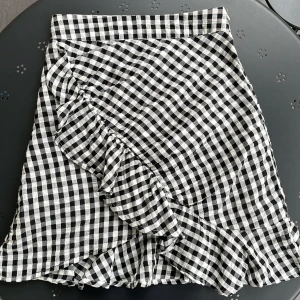 Svartvit rutig kjol - En svartvit rutig kjol med volangdetaljer och dragkedja i sidan.  Kjolen har även en omlott-design. Liten skada i tyget under dragkedjan (se bild 3) men det är inget som syns eller påverkar när man har den på sig 