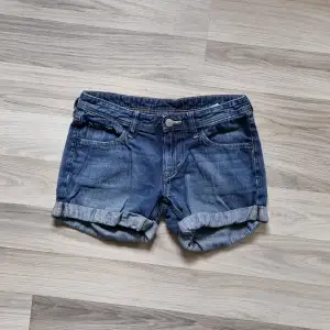 Ett par blå jeansshorts med upprullade ben. Shortsen har fram- och bakfickor samt en knapp och dragkedja framtill.