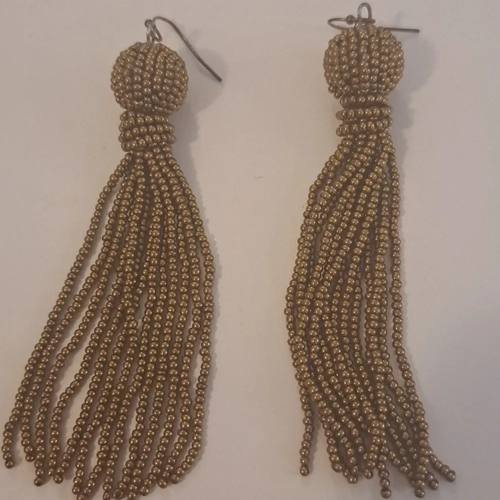 Ett par eleganta örhängen dekorerade med små guldfärgade pärlor. Örhängena har en rund pärlad topp och långa fransar som hänger ner, vilket ger en stilfull och festlig look.. Accessoarer.