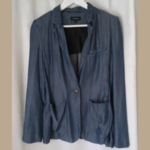 Denimblå mjuk kavaj i lyocell, stl 36 S, blazer, blå, stilren, klassisk - Denimblå kavaj i mjuk och följsam lyocell. En sköldpaddsfärgad knapp fram, knappar i ärmsluten, tre framfickor, tunna axelvaddar. Mått i cm: bröstvidd 44; axelbredd 42; längd fram från axeln 61; ärmlängd 59.