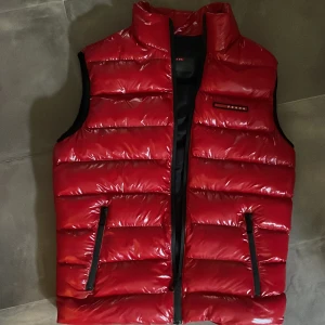 Prada röd väst - Säljer en exklusiv och elegant röd väst från Prada. Västen är i utmärkt skick och har använts sparsamt. Perfekt för lager på lager under kyliga dagar eller som ett stilrent komplement till din outfit. Passar både casual och mer uppklädda tillfällen.