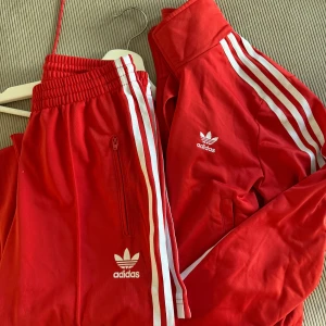 Adidas tracksuit - Oanvänd tracksuit från adidas i den klassiska röda färgen. Tröjan har krage och byxorna är raka/utsvängda med dragkedja!
