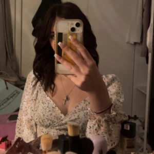 Vit blommig blus - En vit blommig blus med långa ärmar och volangdetaljer vid ärmsluten. Blusen har ett omlottmönster och är tillverkad i ett lätt och luftigt material.