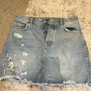 Jeanskjol med slitningar - Säljer en cool jeanskjol med slitningar och knappar framtill. Kjolen är i ljusblå denim och har en rå kant nedtill som ger en avslappnad look. Perfekt för sommarens alla äventyr! 🥰