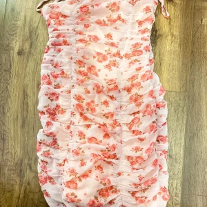 Rosa blommig klänning  - Kort rosa blommig klänning med snören som man knyter vid axeln med och dragkedja för att stänga klänningen. Klänningen har använts 1 gång tidigare.