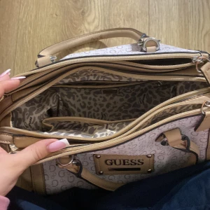 Beige handväska från Guess - Säljer en snygg beige handväska från Guess. Väskan har ett mönster med små G-logotyper och detaljer i ljusbrunt skinn. Den har flera fack inuti och en dragkedja för att hålla dina saker säkra. Perfekt för både vardag och fest!