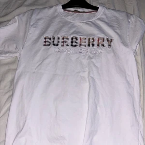 Vit t-shirt från Burberry - Säljer en snygg vit t-shirt från Burberry med texten 'BURBERRY LONDON ENGLAND' på bröstet. T-shirten är i storlek M och har korta ärmar. Perfekt för en casual look eller att klä upp med en snygg jacka. Passar till alla tillfällen!