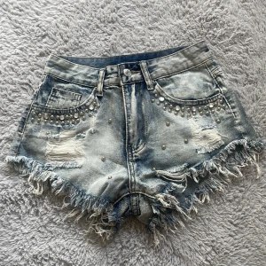 Jeansshorts med pärlor och slitningar - Säljer ett par snygga helt oanvända jeansshorts i storlek XS. De är ljusblå med coola slitningar och pärldetaljer både fram och bak. Shortsen har fransiga kanter och en låg midja. Perfekta för sommaren och festivaler! 🩷👌