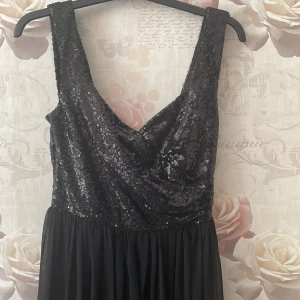 Svart långklänning med paljetter - Säljer en superfin svart långklänning med paljetter upptill. Klänningen har en djup v-ringning och är ärmlös, vilket gör den perfekt för fest eller bal. Kjoldelen är flödande och ger en elegant look. Den är i nyskick och redo att bäras till nästa stora event! Använd en gång, säljer då den inte kommer till användning mer. Köparen står för frakten!
