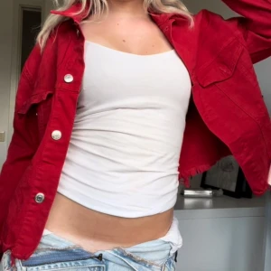 Röd jeansjacka - Säljer en snygg röd jeansjacka. Den har en cool och avslappnad stil med silverfärgade knappar och två bröstfickor. Perfekt för vår och sommar! Jackan är croppad och har en normal passform. Passar perfekt till jeans eller kjol för en trendig look.