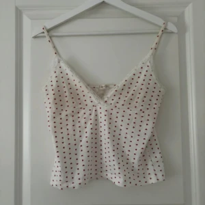 Brandy Melville topp - En söt vit linne topp med små röda hjärtan som mönster. Toppen har smala axelband och spetsdetaljer vid halsringningen.