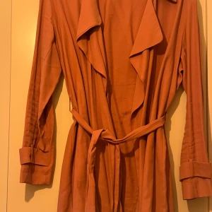 Rostfärgad trenchcoat med knytbälte - Säljer en snygg rostfärgad trenchcoat med knytbälte i midjan. Jackan har långa ärmar med knappar vid ärmsluten och en avslappnad passform. Perfekt för höst och vår! använt nog gg. i bra skick💛🧡