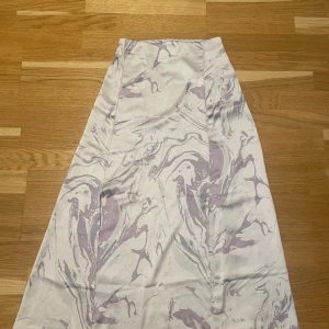 Satin långkjol med mönster - Oanvänd långkjol i satin med två slits fram. Färgen är cremevit med lila mönster. Väldigt skönt material och sitter superfint. Döljd dragkedja i sidan samt resår i midjan. Går ungefär 10/15cm över foten på mig som är 165cm. Köpt för 450kr
