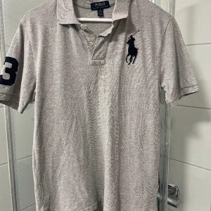 Ralph lauren pike  - En grå Ralph Lauren pike i grymt bra skick. Märkena på bröstet och på högra armen är inbeordrade med en väldigt mörkblå tråd. 