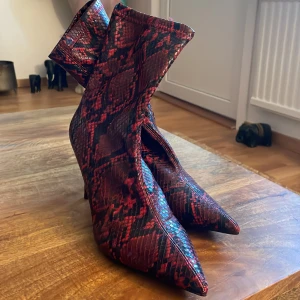 Röda ormskinnsmönstrade stövletter från MNG - Ett par röda stövletter med ormskinnsmönster från MNG. Skorna har spetsig tå och hög klack, vilket ger dem en elegant och iögonfallande look.