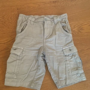 Beige cargo shorts - Beige cargo shorts med flera fickor, inklusive två framfickor, två bakfickor med knappar och två sidofickor. Shortsens midja är elastisk och de har en knapp och dragkedja framtill.