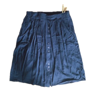 Blå kjol - Behövs strykas i bilden. Väldigt tunn med insydda vikningar, bara knappar på framsidan.   Om högmiddjad går den ner till precis under mina knän (175 cm) Om lågmidjad går den till mitten av mina vader.  (Mått kommer efter förfrågan)  