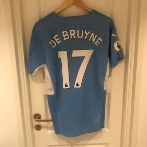  Manchester City fotbollströja 2021/22 HemmaStäll - En blå Manchester City fotbollströja från Puma med korta ärmar. Tröjan har 'De Bruyne' och nummer 17 tryckt på baksidan. På framsidan finns Manchester Citys klubbmärke, Puma-logotypen, och sponsorn 'Etihad Airways'. Tröjan har även Premier League-logotypen på ärmen.