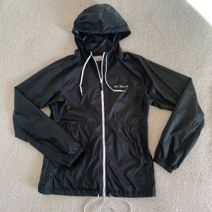 Columbia vindjacka / vårjacka / träningsjacka - Svart vindjacka från Columbia med deras klassiska logga på bröstet. Aldrig använd, utan anmärkningar. Storlek XS men upplevs väldigt rymlig och passar även S utan problem. Tunn jacka med fickor och luva med snören även vid midjan.