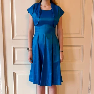 Blå klänning med kort kofta, Emmy Design - Jättefin blå klänning med en kort kofta i skönt material. Storlek 38.  Modellen på bilden är 173 cm och har storlek 38 i de flesta andra kläder. Aldrig använd. Vi har lagt ner vår klänningsbutik och säljer ut. Ny pris är 2000-3000kr