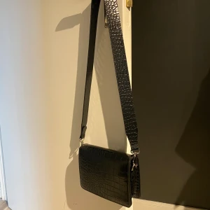Liten handväska  - Liten axelremsväska i krokodilmänster. Väskan är använd ett fåtal gånger och är i nyskick. Den har ett ytterfack på baksidan av väskan och ett mindre dragkedjefack på insidan. Köpt på Lindex förra året för 299kr. Pris kan diskuteras💕