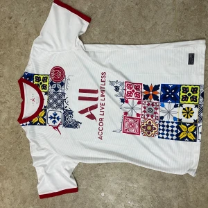 PSG x Jordan vit kortärmad fotbollströja - Säljer en vit PSG x Jordan fotbollströja med röda detaljer vid hals och ärmslut. Framsidan har färgglada grafiska mönster i blått, gult och rött samt PSG-logga och Accor Live Limitless-tryck. Tröjan är tillverkad i lätt och ventilerande polyester.