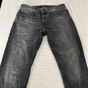 Jack & Jones gråa tapered jeans - Snygga gråa jeans från Jack & Jones i modellen Tapered Mike. Jeansen har en tvättad look med slitningar, klassiska fem fickor och knappgylf. Passformen är tapered med smalare ben och de är tillverkade i jeansmaterial.