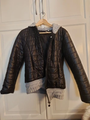 Svart quiltad pufferjacka med stickad huva - ⚠️Det är litet hål i höger ficka!⚠️ Svart pufferjacka med quiltad yta, 2 fram fickor och dragkedja framtill. Jackan har en grå, grovstickad huva och matchande stickad kant nedtill, vilket ger en unik och cool look. Perfekt för dig som gillar att sticka ut med detaljer. Långärmad och med rundad form. 