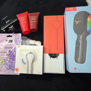 En särpräglad samling - Lyckolådan 🤗: Innehåller en rosa smartklocka, vita Bluetooth-hörlurar, en röd mikrofon, en ansiktsmask, en ring, en ansiktsskrubb och schampo. Och allt är helt nytt!