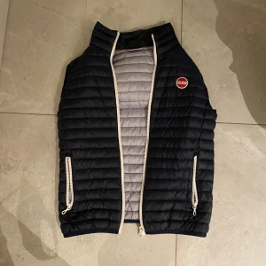 Svart dunväst från Colmar Originals - Snygg svart dunväst från Colmar Originals med quiltad design och ljusgrå insida. Västen har två fickor med vita dragkedjor och ett broderat Colmar-märke på bröstet. Perfekt att slänga över en hoodie för extra värme. Ståkrage och stilren look.