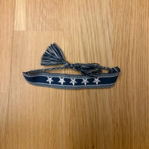 Blå tygarmband med vita stjärnor - Snyggt blått armband i tyg med vita broderade stjärnor längs bandet. Armbandet har knytning med två tofsar i ändarna för en cool och avslappnad look. Perfekt accessoar för att ge din outfit lite extra edge. Finns även i svart om man vill 🤩