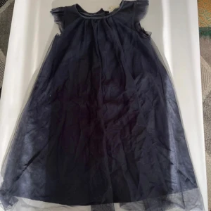 Mörkblå tyllklänning från H&M - Supersöt mörkblå klänning från H&M med tyllöverdrag och lätt puffiga axlar. Klänningen har en vid, luftig passform och är ärmlös, perfekt för dig som gillar en klassisk look med lite extra volym.