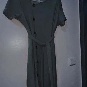 Ljusgrön klänning med knappar från SHEIN - Snygg ljusgrön klänning från SHEIN Curve med knappar framtill och matchande bälte i midjan. Klänningen har korta ärmar och en rak, lång siluett. Perfekt för dig som gillar enkel men stilren design.