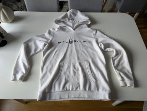 Vit hoodie från Sail Racing - Vit hoodie från Sail Racing med svart logga på bröstet. Klassisk känguruficka framtill, huva och långa ärmar. Perfekt för en chill och sportig stil. Mjuk och skön bomullskänsla.
