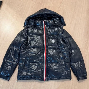 Svart Moncler pufferjacka med huva - Svart pufferjacka från Moncler med glansig finish och huva. Jackan har en röd, vit och blå dragkedja, Moncler-logga på bröstfickan och flera praktiska fickor. Fylld med certifierat gåsdun och har coolt serietryck på insidan. Size M