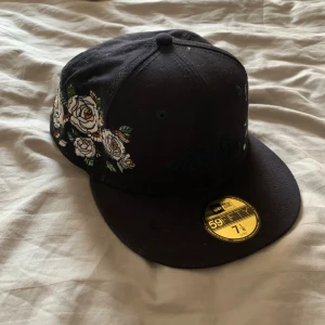 Limiterad New Era Keps - Snygg svart New Era 59FIFTY keps med broderade vita rosor och gröna blad på sidorna. Platt skärm med grön undersida och klassisk gul etikett framtill. Perfekt för dig som vill sticka ut med en unik och limiterad keps.