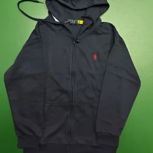 Mörkblå hoodie från Polo Ralph Lauren - Snygg mörkblå hoodie från Polo Ralph Lauren med dragkedja framtill och klassisk röd logobrodering på bröstet. Tröjan har huva med snörning, två fickor och ribbade muddar vid ärmslut och nederkant. Perfekt för chill dagar och streetstyle.