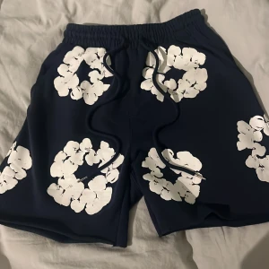 Blå blommiga shorts från Denim Tears - Säljer ett par mörkblå shorts från Denim Tears med stora vita blommönster över hela plagget. Shortsen har elastisk midja med snörning och en bakficka. Perfekta för sommaren och ger en chill vibe. Perfekt för dig som vill bli få en glowup