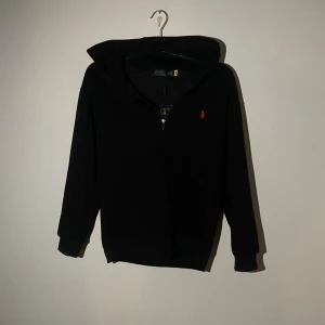 Ralph Lauren Hoodie - Svart hoodie från Polo Ralph Lauren med röd broderad logga på bröstet. Tröjan har huva, dragkedja framtill och långa ärmar med ribbade muddar. Tillverkad i mjuk bomull som är skön att ha på sig. Tröjan är oanvänd och är redo för en användare. Perfekt för dessa kalla dagar!!🙌