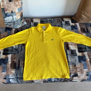 Gul långärmad pikétröja från Lacoste - Snygg gul pikétröja från Lacoste med klassisk krage och två vita knappar framtill. Tröjan har lång ärm och det ikoniska krokodilmärket broderat på bröstet. Perfekt för dig som vill sticka ut med färg och stil.