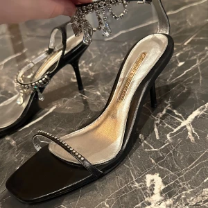 Svarta klack-sandaler Buffalo London - Säljer ett par svarta sandaler med klack från Buffalo London. Skorna har en fyrkantig tå, silverfärgad innersula och smala remmar dekorerade med strass. Runt ankeln sitter en glittrig kedja med hängande kristaller som ger extra bling.