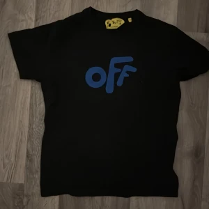 Svart Off-White t-shirt med blå tryck - Svart t-shirt från Off-White med blått 'OFF' tryck på framsidan och ikoniska blå pilar på ryggen. Klassisk rund hals och korta ärmar. Materialet är mjuk bomull och har en gul etikett i nacken med Off-White Kids branding. Perfekt för dig som gillar streetwear.