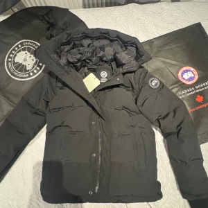  Canada Goose Macmillan  - Svart pufferjacka från Canada Goose med ikonisk Arctic Program patch på ärmen. Jackan har huva, dragkedja framtill och två stora fickor. Fodrad för extra värme och tillverkad i slitstarkt syntetmaterial. Perfekt för kalla vinterdagar och har en clean, modern look. Den är i storlek m och är ny 