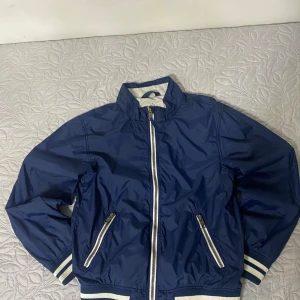 Blå vindjacka från H&M L.O.G.G. - Snygg marinblå vindjacka från H&M L.O.G.G. med vita detaljer och dragkedjor. Jackan har ribbade muddar med ränder, ståkrage och två fickor med dragkedja. Materialet är polyamid och polyester med inslag av bomull. Perfekt för blåsiga dagar.