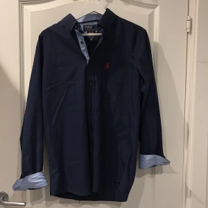 Mörkblå skjorta Polo Ralph Lauren - Snygg mörkblå skjorta från Polo Ralph Lauren i slim fit-modell. Skjortan har klassisk krage, knappar framtill och ett rött broderat Polo-logo på bröstet. 