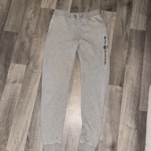 Grå mjukisbyxor från Sail Racing - Grå mjukisbyxor från Sail Racing med svart logga längs benet. Byxorna har snörning i midjan och ribbade muddar vid bensluten. Tillverkade i mjuk bomullsblandning för maximal komfort. Perfekta för chill eller träning. XS- S