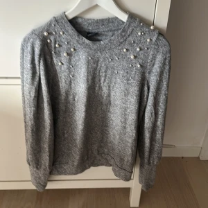 Grå tröja med pärldetaljer från Gina Tricot - Superfin grå tröja från Gina Tricot med pärlor på axlar och över bröstet. Mjuk och skön med lång ärm och rund halsringning. Perfekt för dig som vill ha något extra till din outfit!